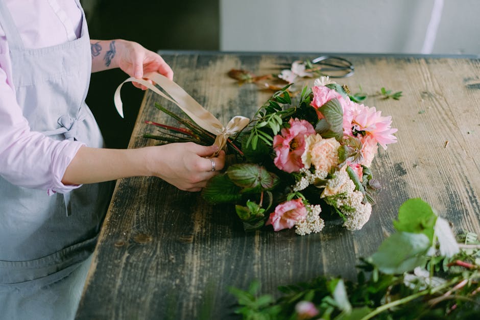 A pair of hands arranging a floral bouquet on a dark workspace surface, featuring light pink, white, and yellow roses, along with green eucalyptus and lily leaves. The person is holding a spool of twi