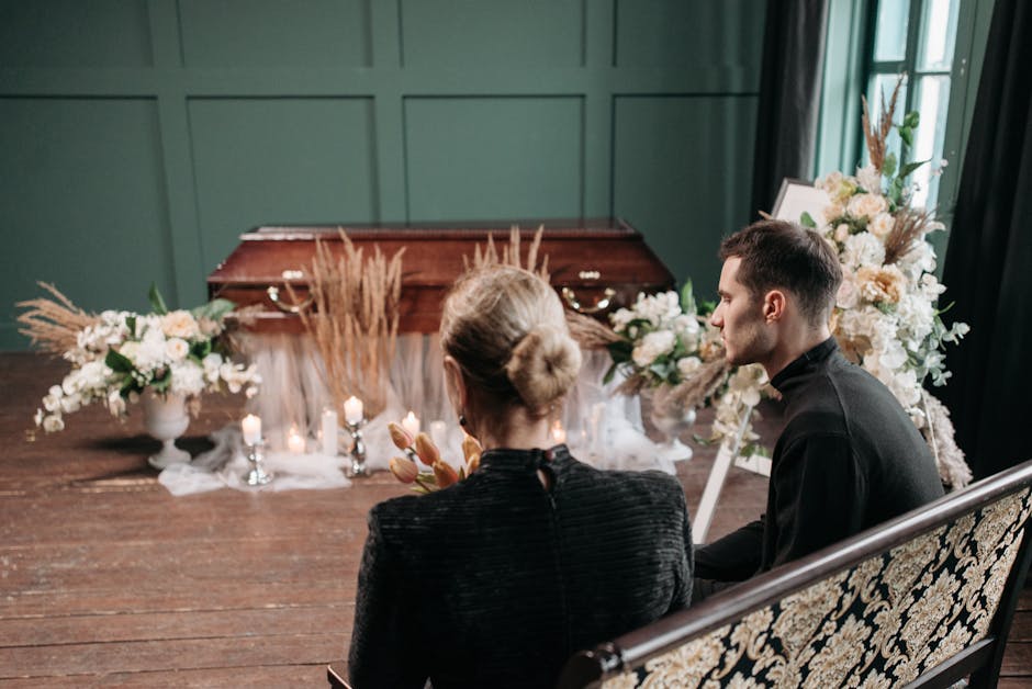 A woman seated in a vintage floral-patterned armchair holding a bouquet of pastel-colored flowers, with her eyes closed in a moment of reflection. In front of her, a wooden casket draped with white tu