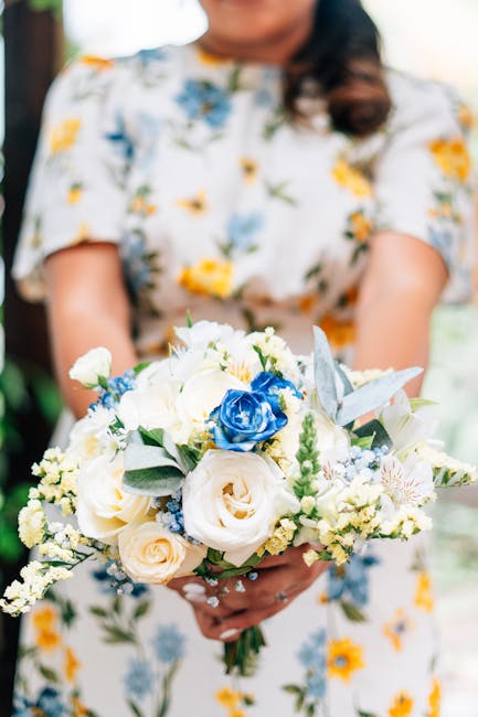 A wedding scene featuring a couple sharing a kiss in front of a floral arch adorned with red, white, and pink flowers, with their hands holding a bouquet of white roses. The groom is dressed in a trad