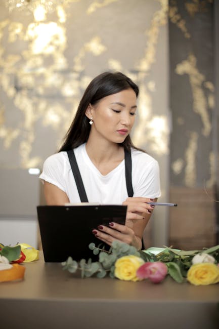 A florist and a customer are engaged in a discussion inside a flower shop. The florist, dressed in a white sleeveless top and beige apron, stands beside a wooden table covered with pink roses, white l