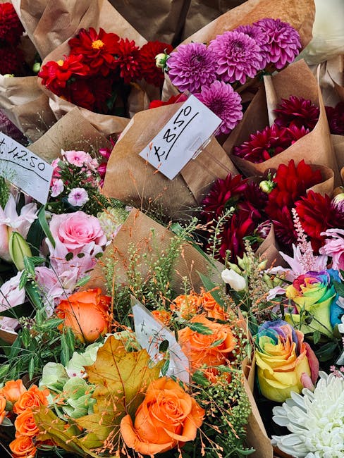Exterior display of multiple plastic crates filled with potted flowers, prominently featuring vibrant pink, red, white, yellow, and orange blooms. The flowers include carnations and chrysanthemums, wi