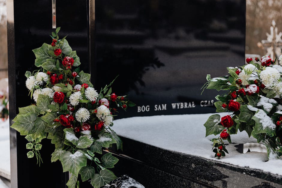 A close-up view of a black marble funeral memorial adorned with floral tributes, featuring arrangements of red roses, white chrysanthemums, and green ivy leaves. The flowers are freshly cut, with the 