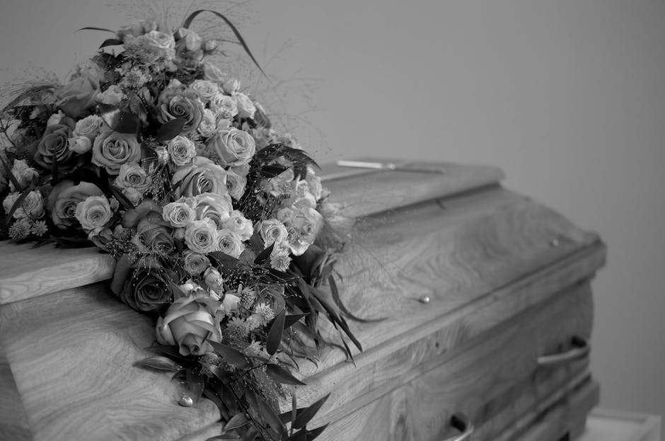 A close-up view of a black marble funeral memorial adorned with floral tributes, featuring arrangements of red roses, white chrysanthemums, and green ivy leaves. The flowers are freshly cut, with the 