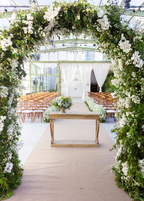 A floral arrangement featuring a large, semi-circular arch densely decorated with white and green flowers, including baby's breath and greenery, set against a textured stone wall. Beneath the arch, a 